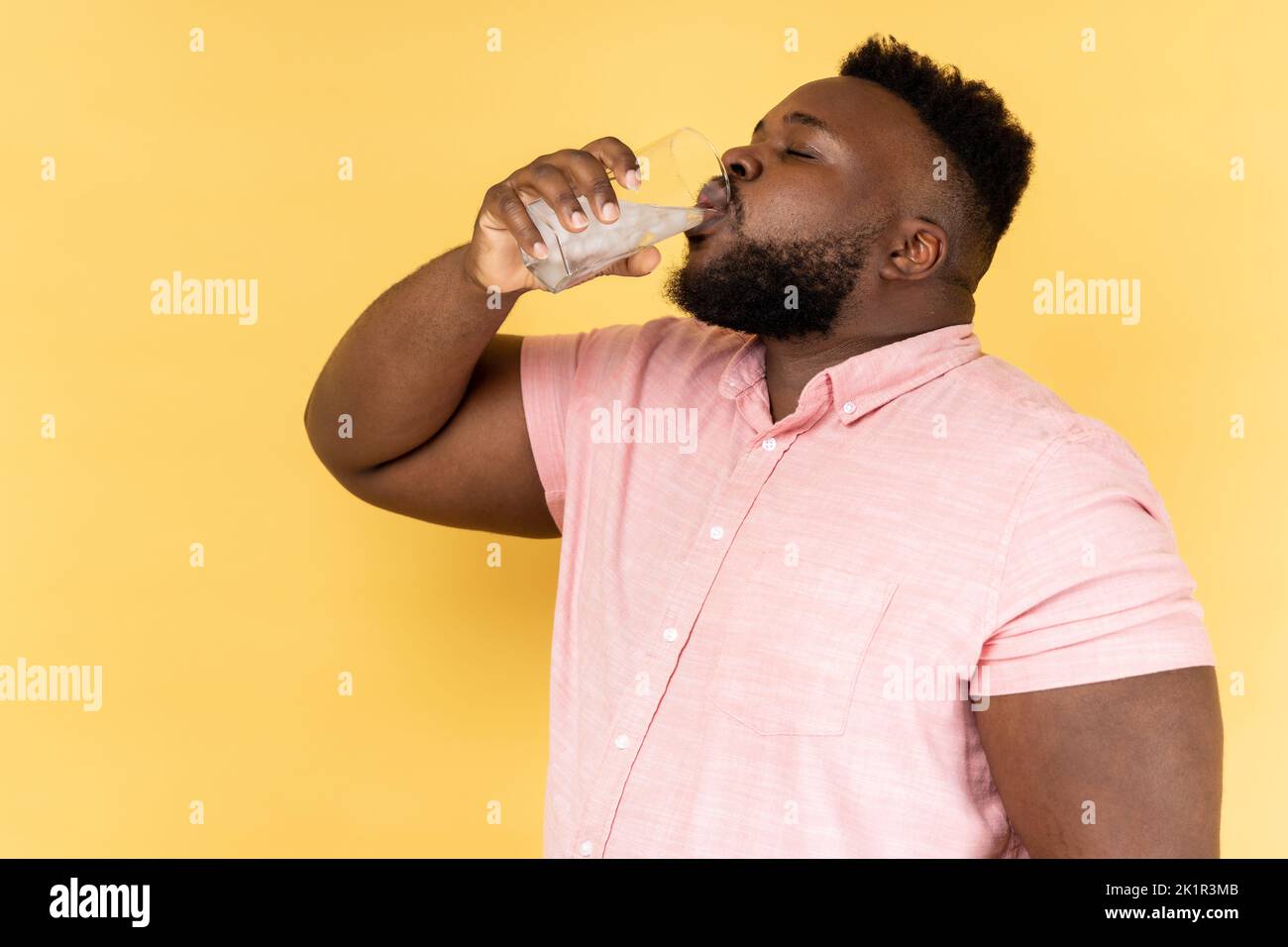 Vista laterale dell'uomo portatore con camicia rosa acqua potabile con ghiaccio dal vetro, si sente assetato, gustando bevande fredde durante le calde giornate estive. Studio al coperto isolato su sfondo giallo. Foto Stock