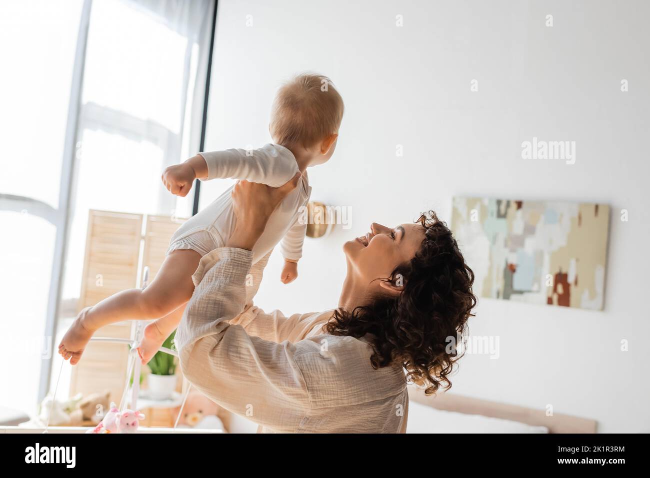 madre felice e ricciola in loungewear tenendo in braccio bambino figlia in romper, immagine di scorta Foto Stock