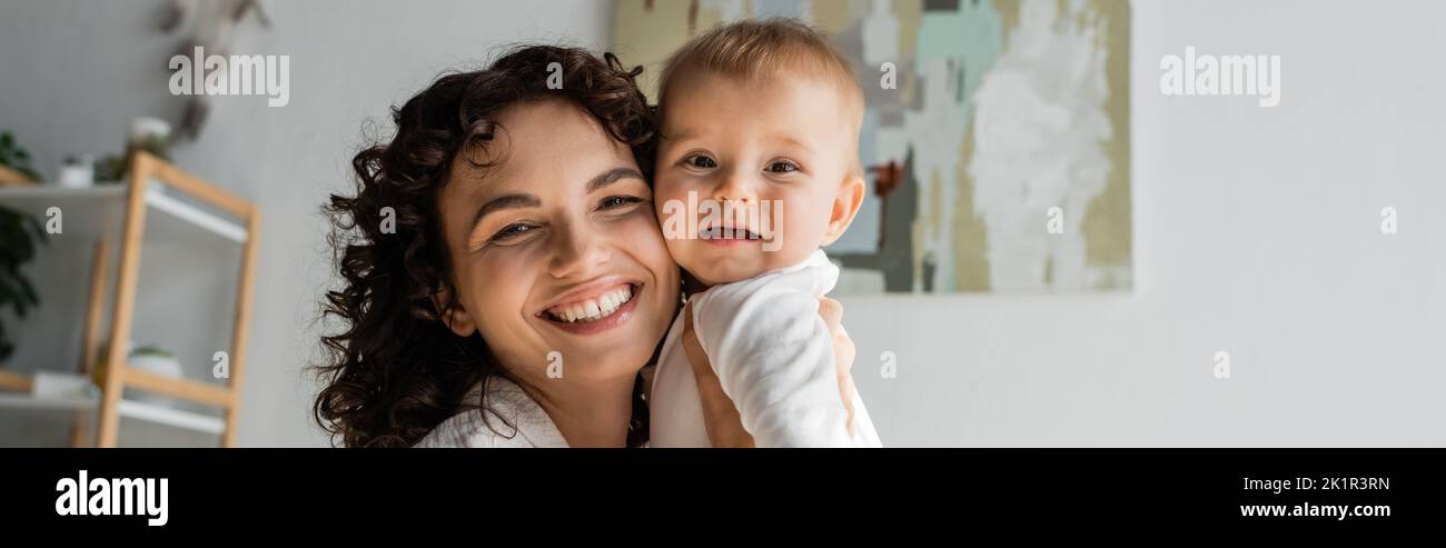 donna felice e curly che tiene in braccia bambino figlia in romper, banner, immagine stock Foto Stock