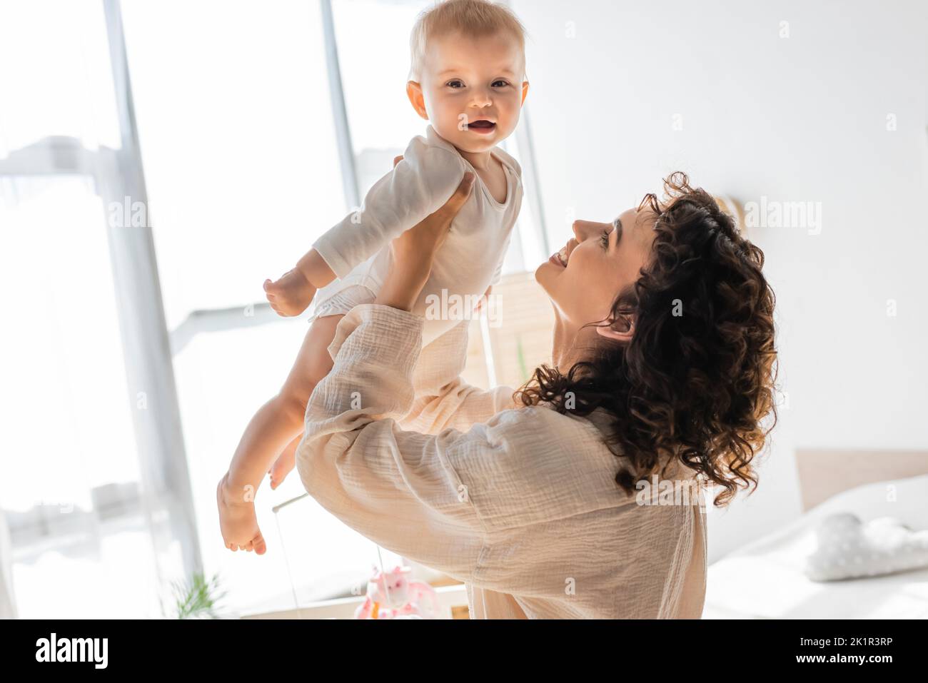 donna felice e riccia in loungewear tenendo in braccio nuda figlia in romper, immagine di scorta Foto Stock