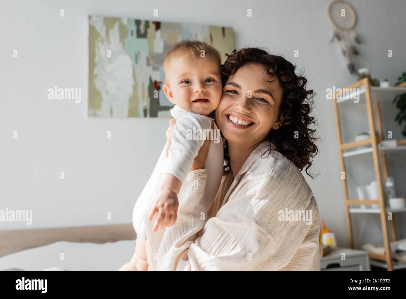 donna felice e ricciola in loungewear tenendo in braccio bambina in romper, immagine di scorta Foto Stock