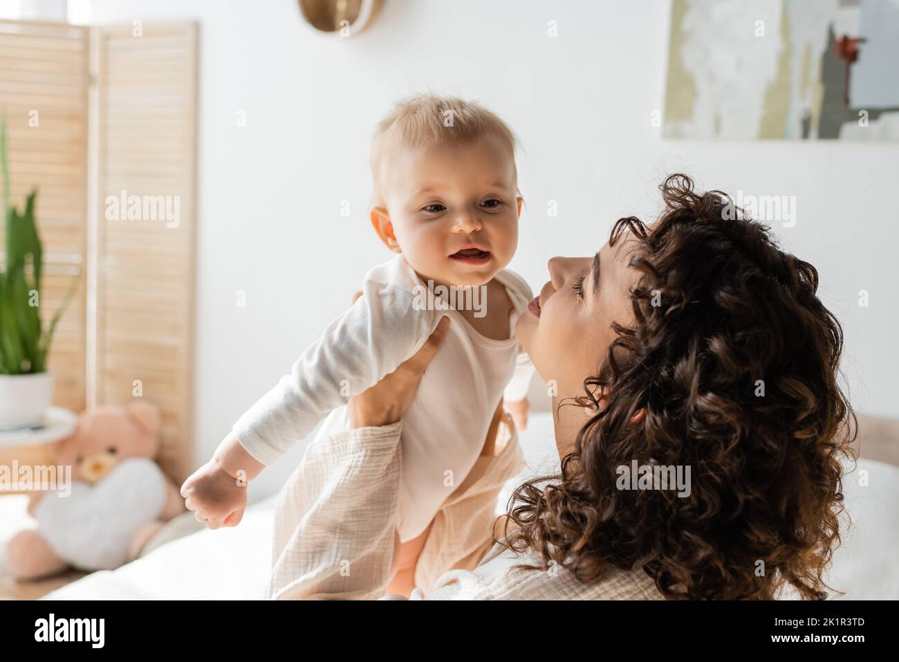 donna felice e riccia in loungewear tenendo in braccio bambino in romper, immagine di scorta Foto Stock