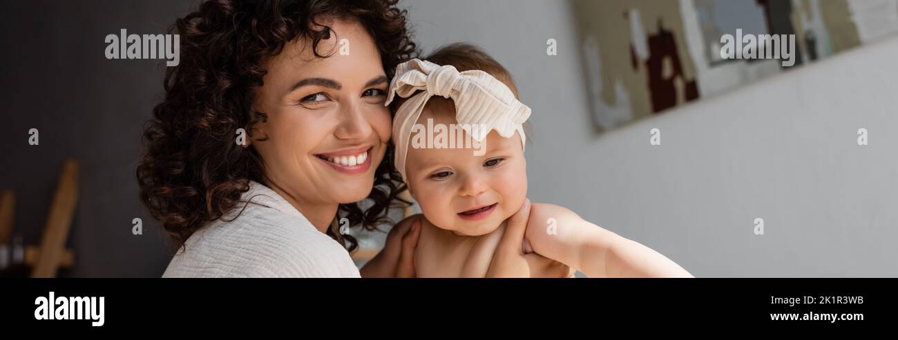allegro e riccio madre che tiene in braccio bambina in fascia con prua, banner, immagine stock Foto Stock