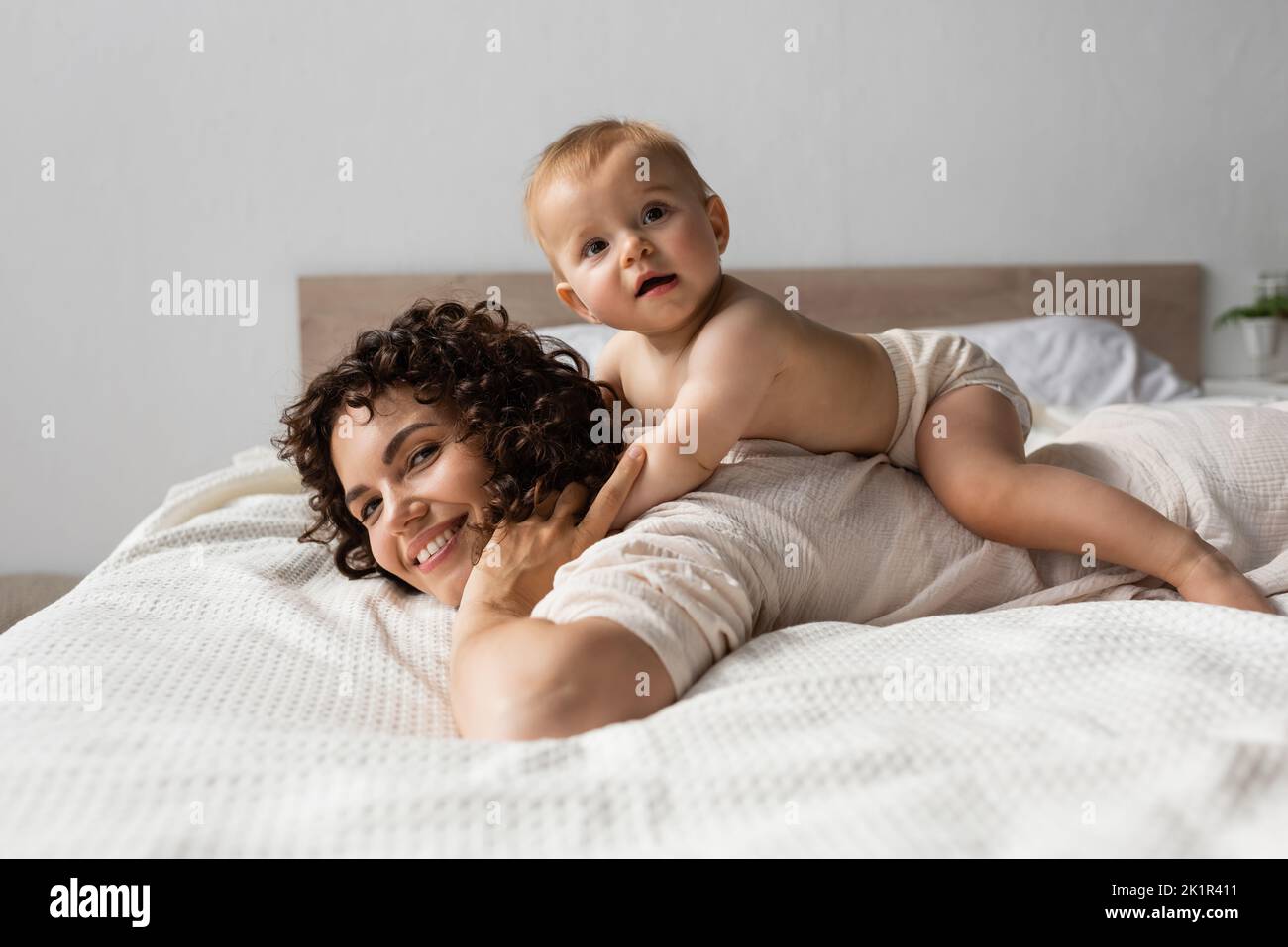 bambino bambino sdraiato sulla parte posteriore della madre felice con i capelli ricci in camera da letto, immagine di scorta Foto Stock