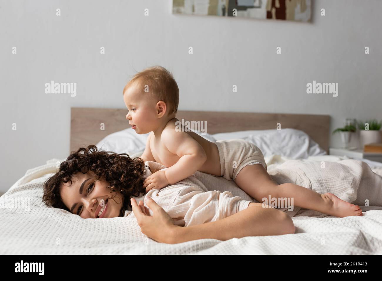 allegro bambino sdraiato sul retro della madre riccia sorridente in camera da letto, immagine stock Foto Stock