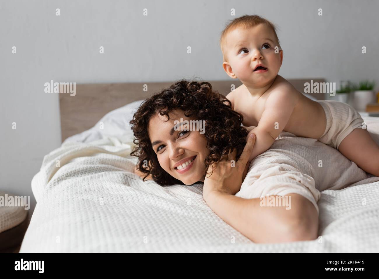 bambino bambino sdraiato sul retro di madre felice con i capelli ricci in camera da letto, immagine stock Foto Stock