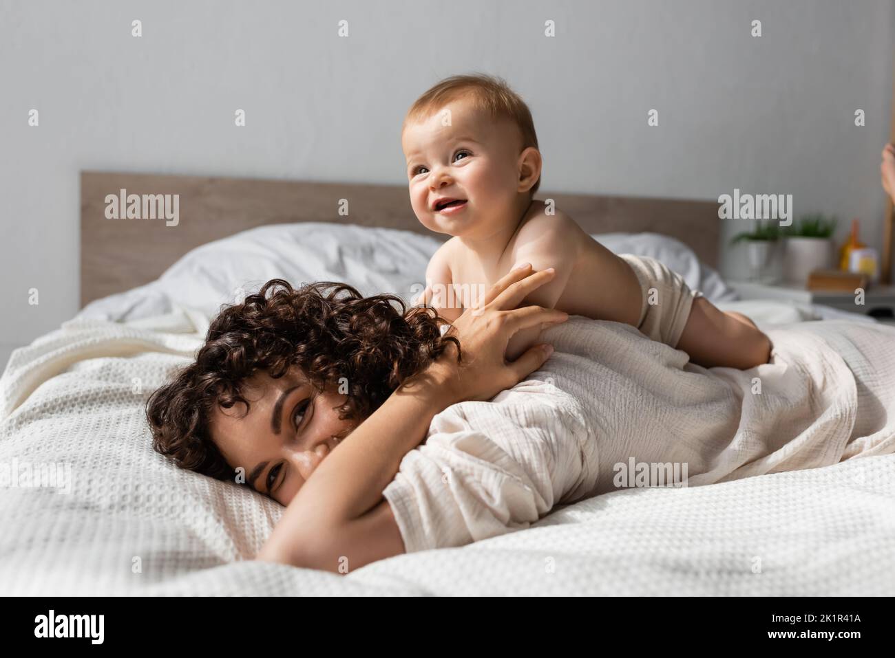 allegro bambino sdraiato sul retro della madre riccio in camera da letto, immagine stock Foto Stock
