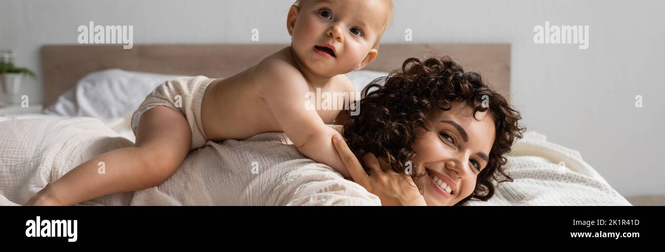 bambino bambino sdraiato sul retro di madre felice con capelli ricci in camera da letto, banner, immagine stock Foto Stock