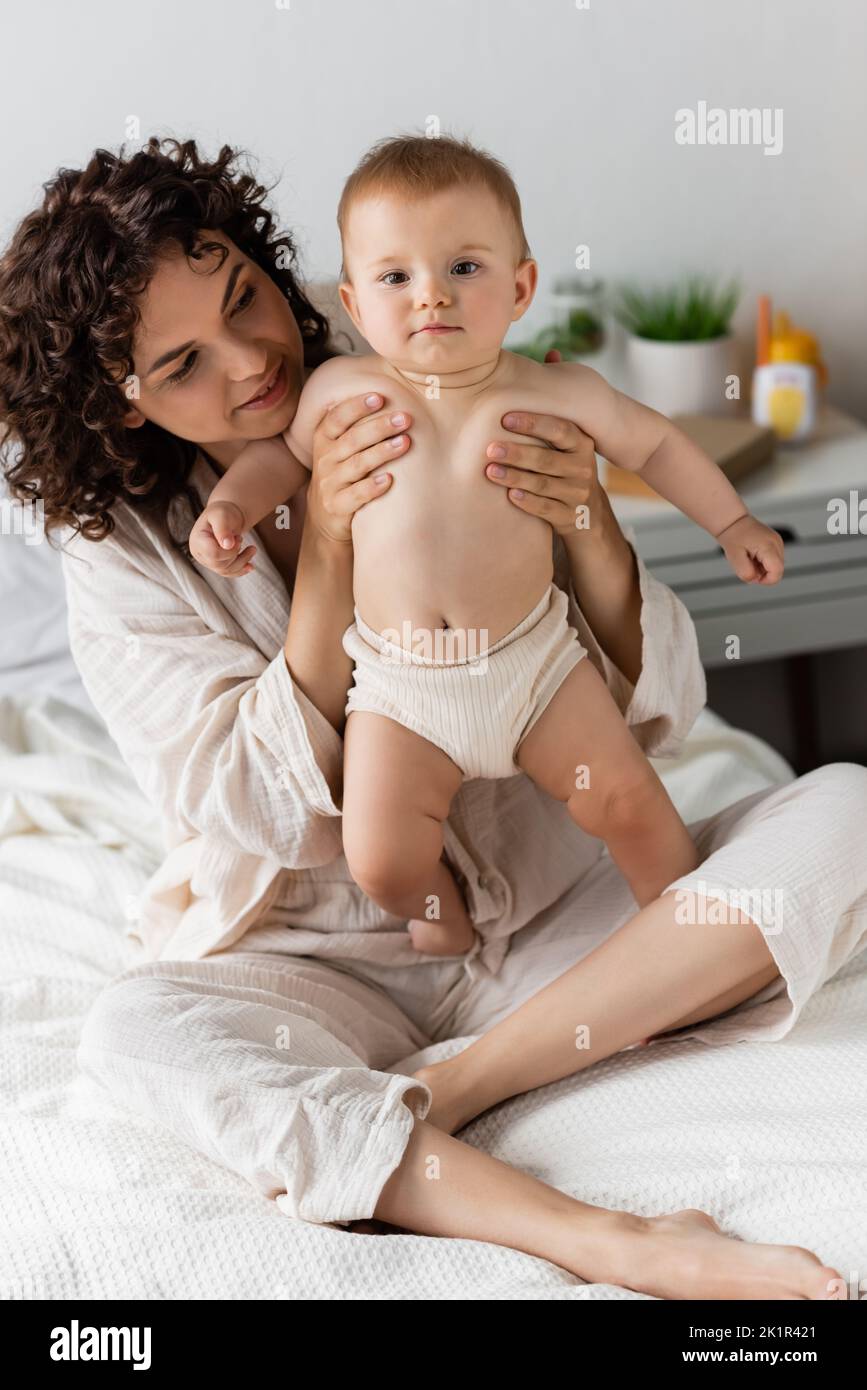 donna compiaciuta con capelli ricci sorridendo mentre tiene in braccio bambino, immagine di scorta Foto Stock
