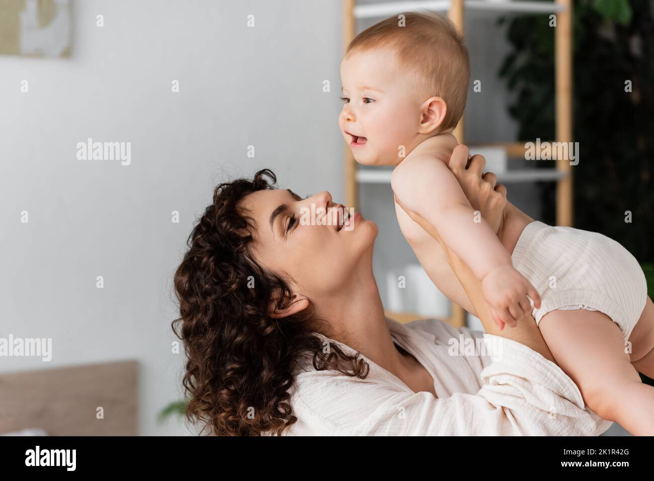 donna compiaciuta con capelli ricci sorridendo mentre tiene in braccio bambino bambino, immagine di scorta Foto Stock