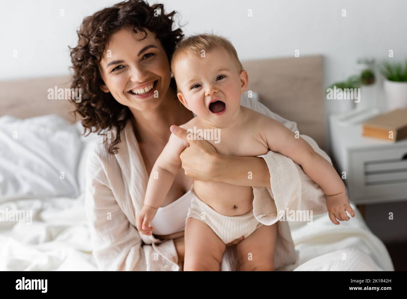 donna allegra in loungewear tenendo in braccio bambino figlia con bocca aperta, immagine stock Foto Stock