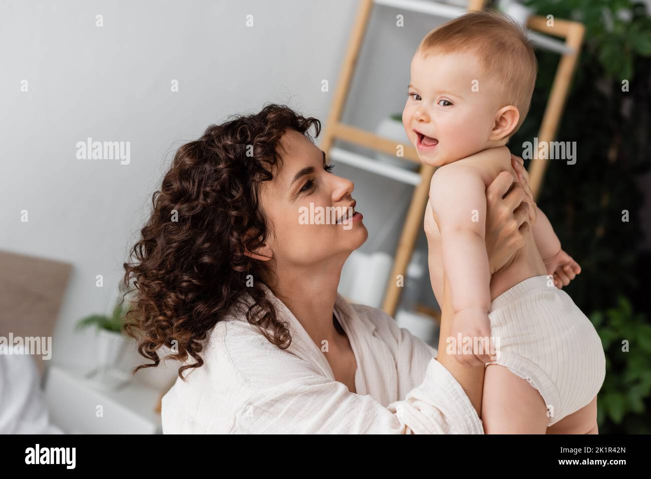 donna compiaciuta con capelli ricci sorridendo mentre tiene in braccio bambino eccitato figlia, immagine di scorta Foto Stock