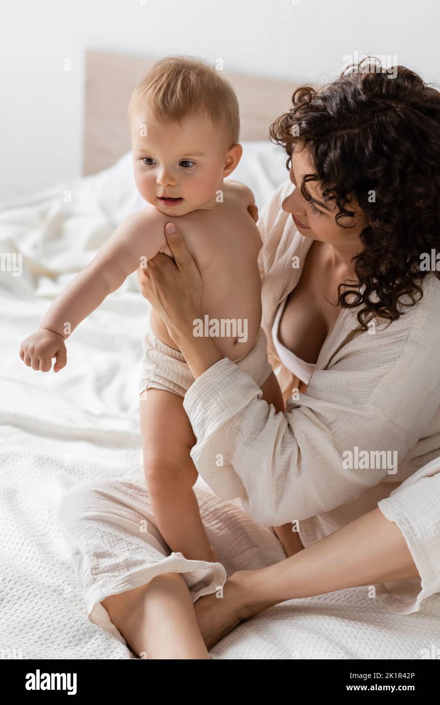 donna con capelli ricci seduta in loungewear a letto e tenendo in braccio bambino figlia, immagine stock Foto Stock