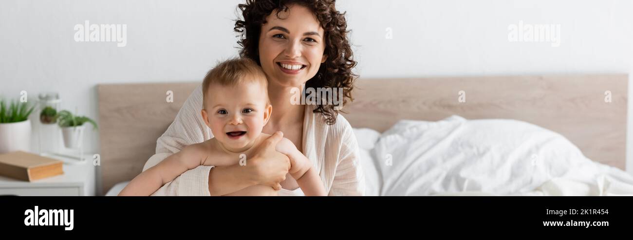 donna gioiosa in loungewear tenendo in braccio sorpresi bambino figlia con bocca aperta, banner, immagine stock Foto Stock