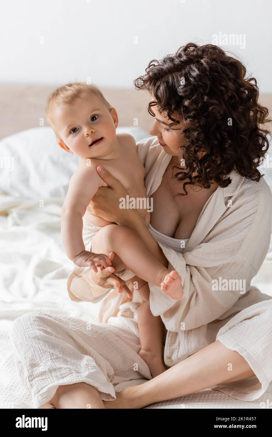 donna con capelli ricci seduta in loungewear a letto e tenendo in braccio felice bambino figlia, immagine stock Foto Stock