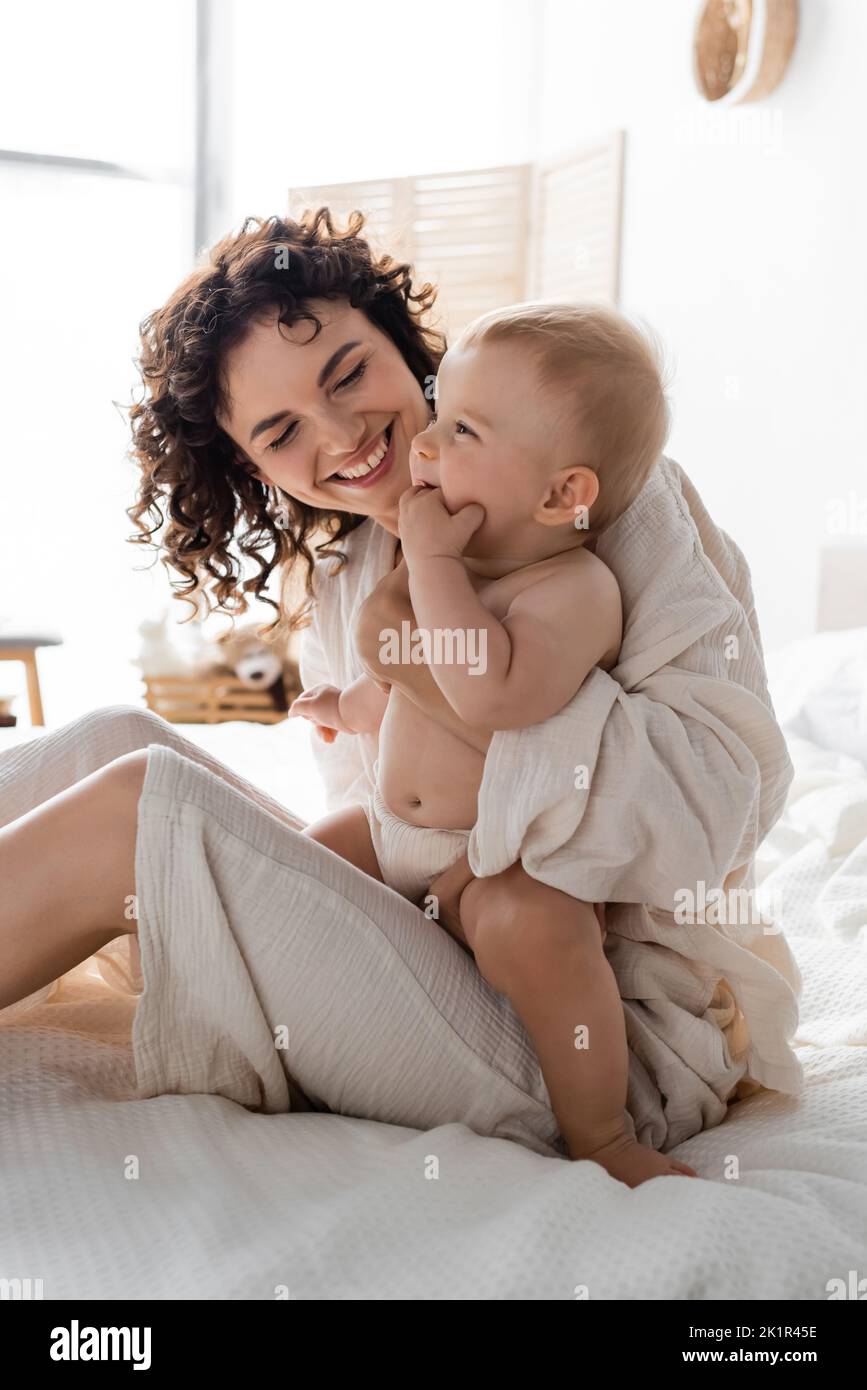donna allegra in loungewear tenendo in braccio bambino figlia e seduta sul letto, immagine stock Foto Stock
