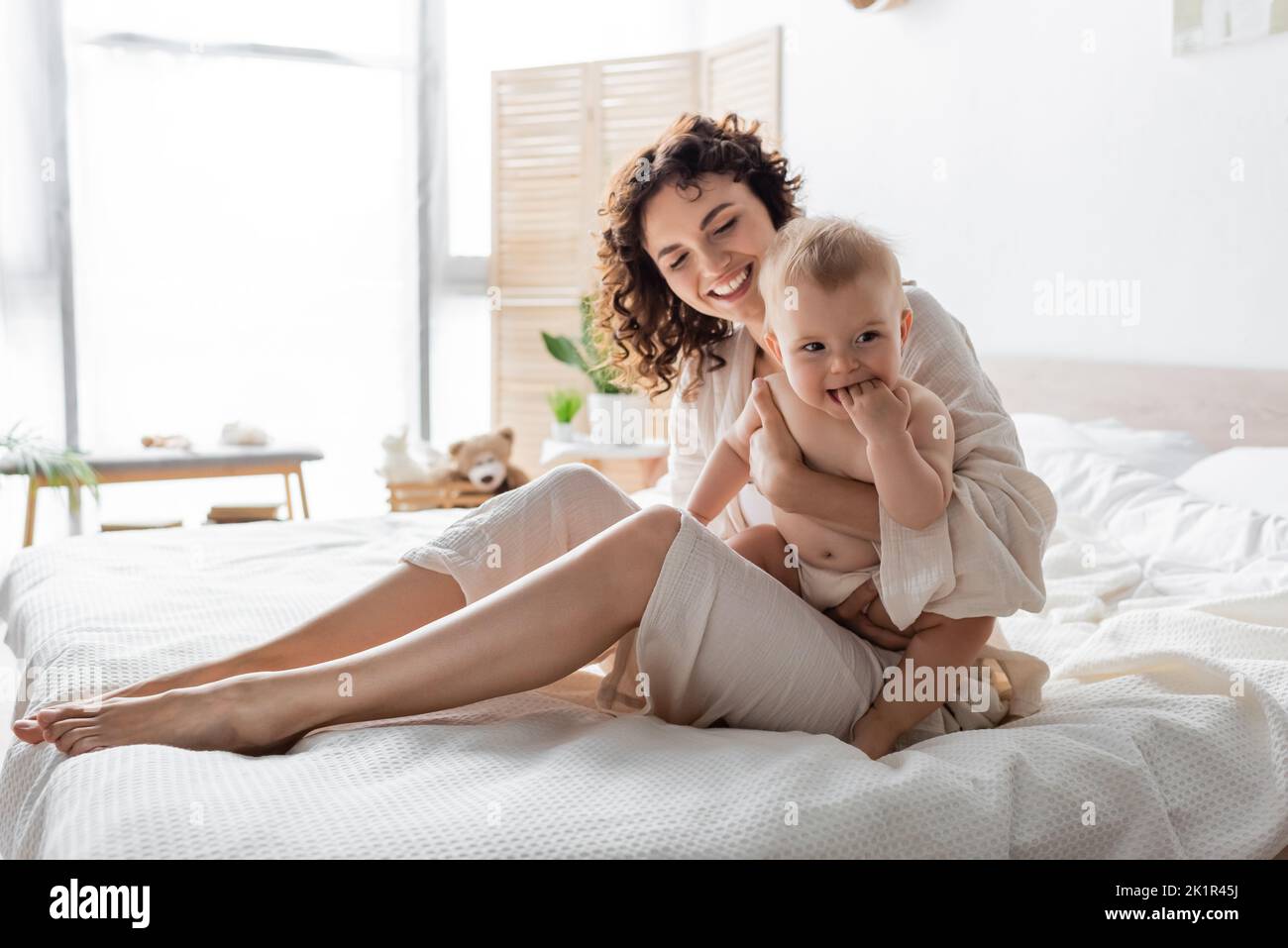 donna allegra in loungewear tenendo in braccio bambino e seduta sul letto, immagine stock Foto Stock