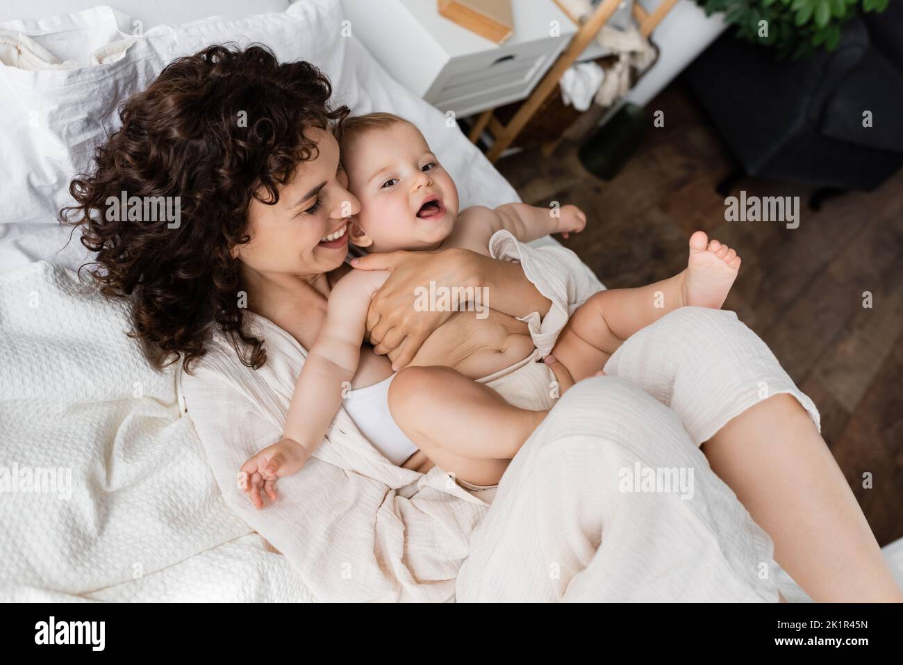 vista dall'alto della donna gioiosa in loungewear tenendo in braccio sorpresi bambino figlia e sdraiato sul letto, immagine stock Foto Stock