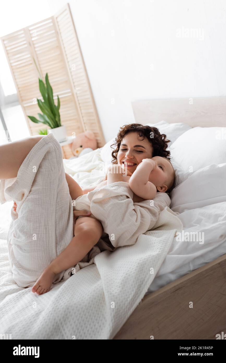 donna allegra in loungewear tenendo in braccio bambino figlia e sdraiato sul letto, immagine stock Foto Stock