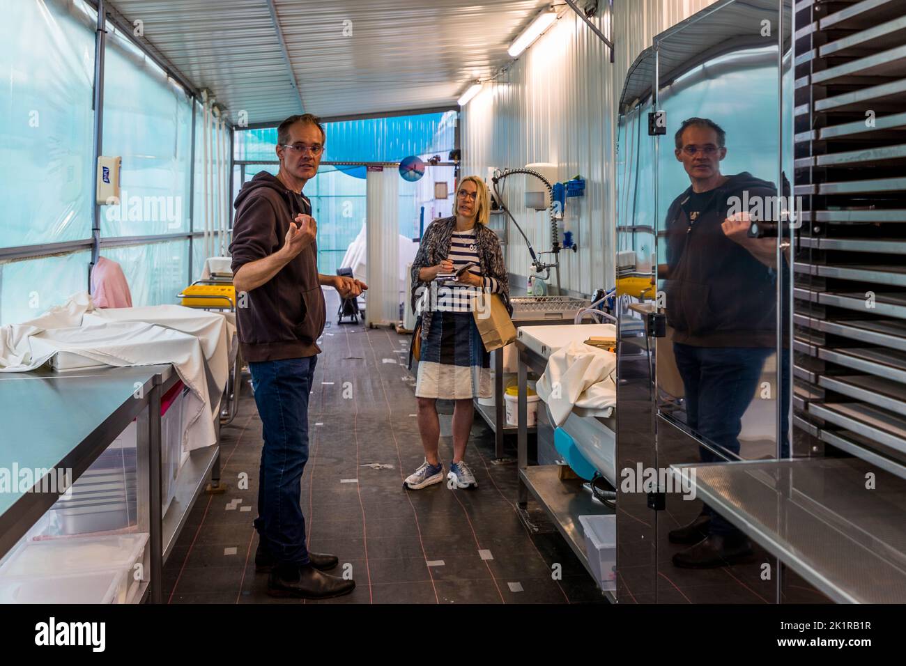 Joël Villeneuve gesticola mentre spiega la sala di lavorazione della sua produzione di spirulina a Saint-Vivien-de-Médoc, Médoc, Francia, mentre la giornalista Angela Berg ascolta di fronte a lui. Uno specchio sulla parete posteriore riflette la scena, con tavoli in acciaio inossidabile e scaffali rivestiti di plastica che costeggiano la stanza stretta. L'azienda familiare EARL Médoc Micro Algues (Spiruline de la Pointe d'Argent) coltiva la spirulina, una cianobatteria nota colloquialmente anche come alghe blu, dal 2018. Foto Stock