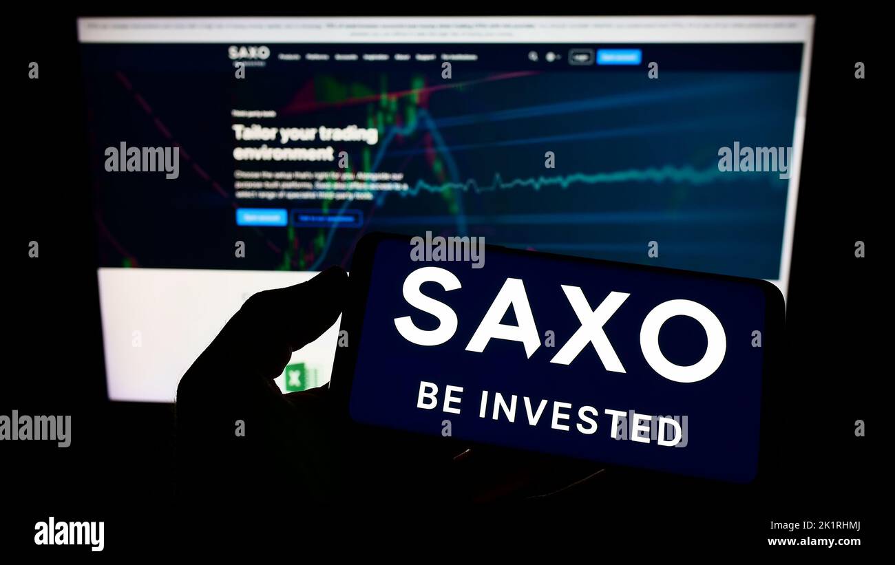Persona che tiene il cellulare con il logo della società danese di banche d'investimento Saxo Bank COME sullo schermo di fronte alla pagina web. Messa a fuoco sul display del telefono. Foto Stock