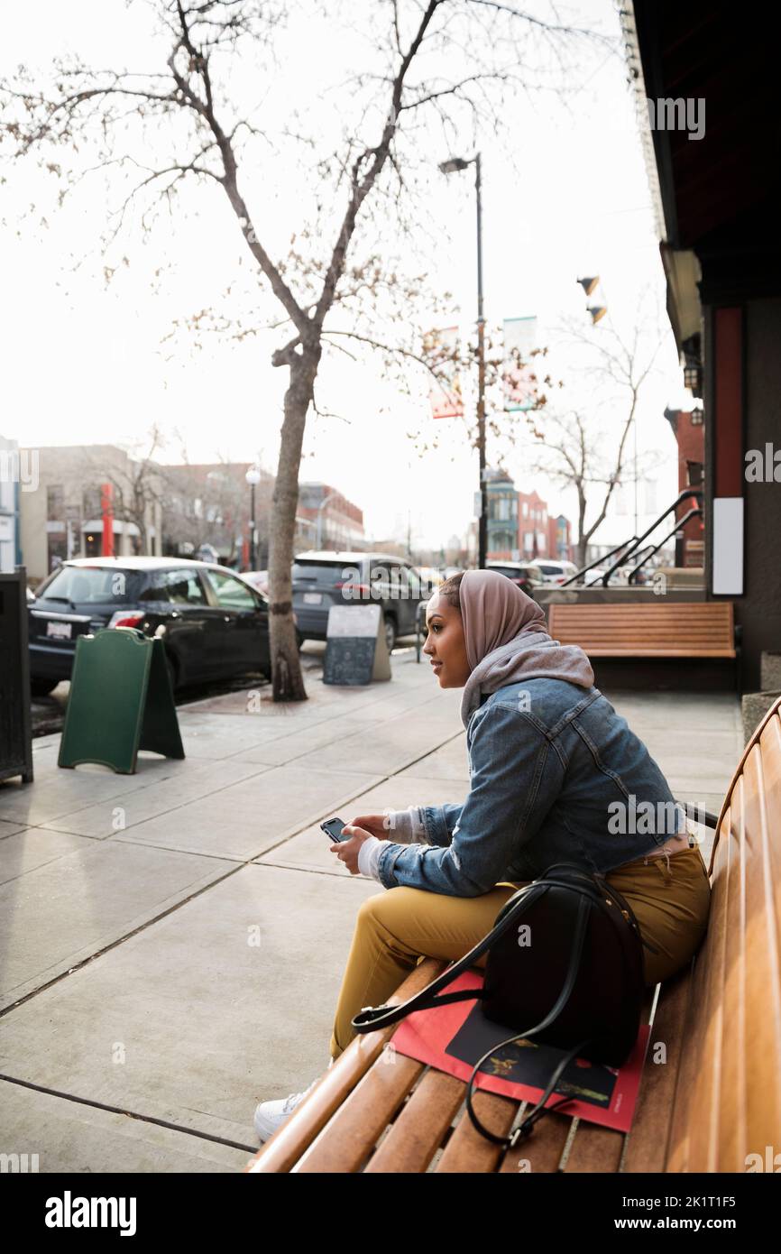Giovane donna con smartphone in attesa sul banco urbano Foto Stock