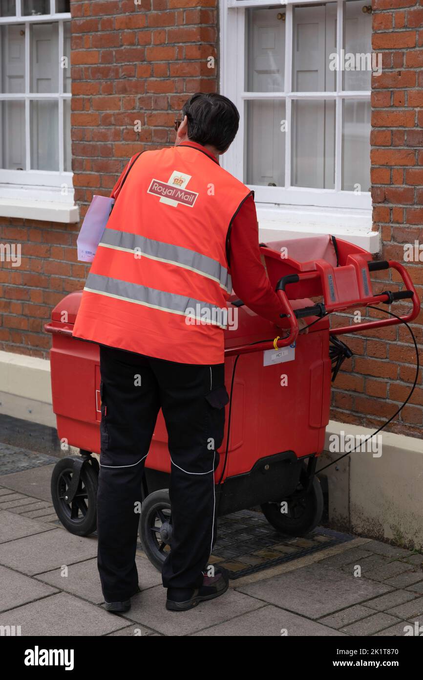 Il postino in giacca arancione impiegato dalla Royal Mail inglese con lettere sotto il braccio chiude il coperchio del carrello pieno di oggetti postali Foto Stock