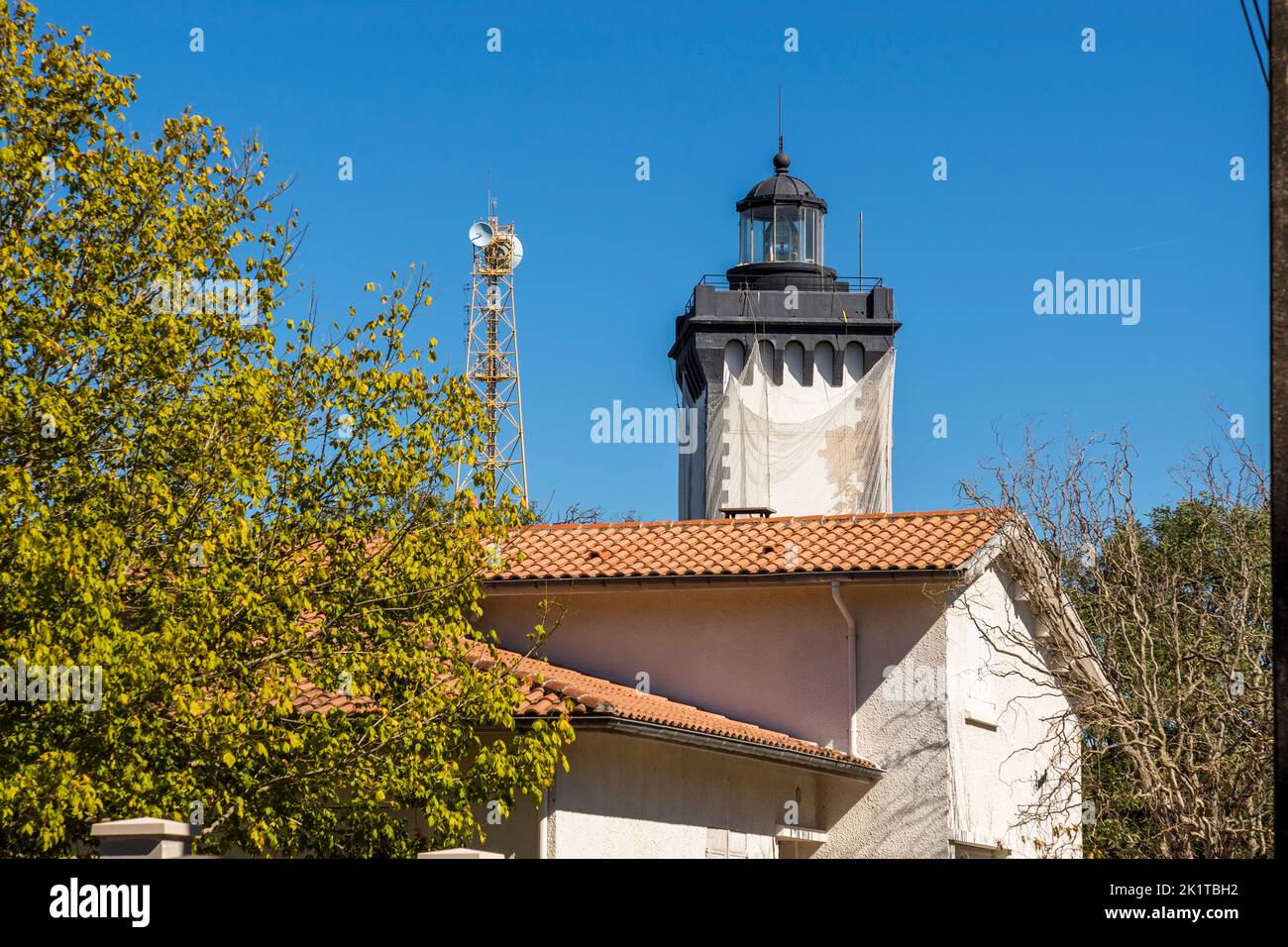 Il faro di Phare de Grave si erge dietro il tetto dell’edificio del museo adiacente vicino a Le Verdon-sur-Mer, Médoc, Francia, avvolto in teli protettivi durante i lavori di ristrutturazione. Automatizzato dal 1955, la torre funge altrimenti da museo per boe e segnali luminosi. Un traliccio con un’antenna di ripetitore radio si trova alla sua sinistra. Foto Stock