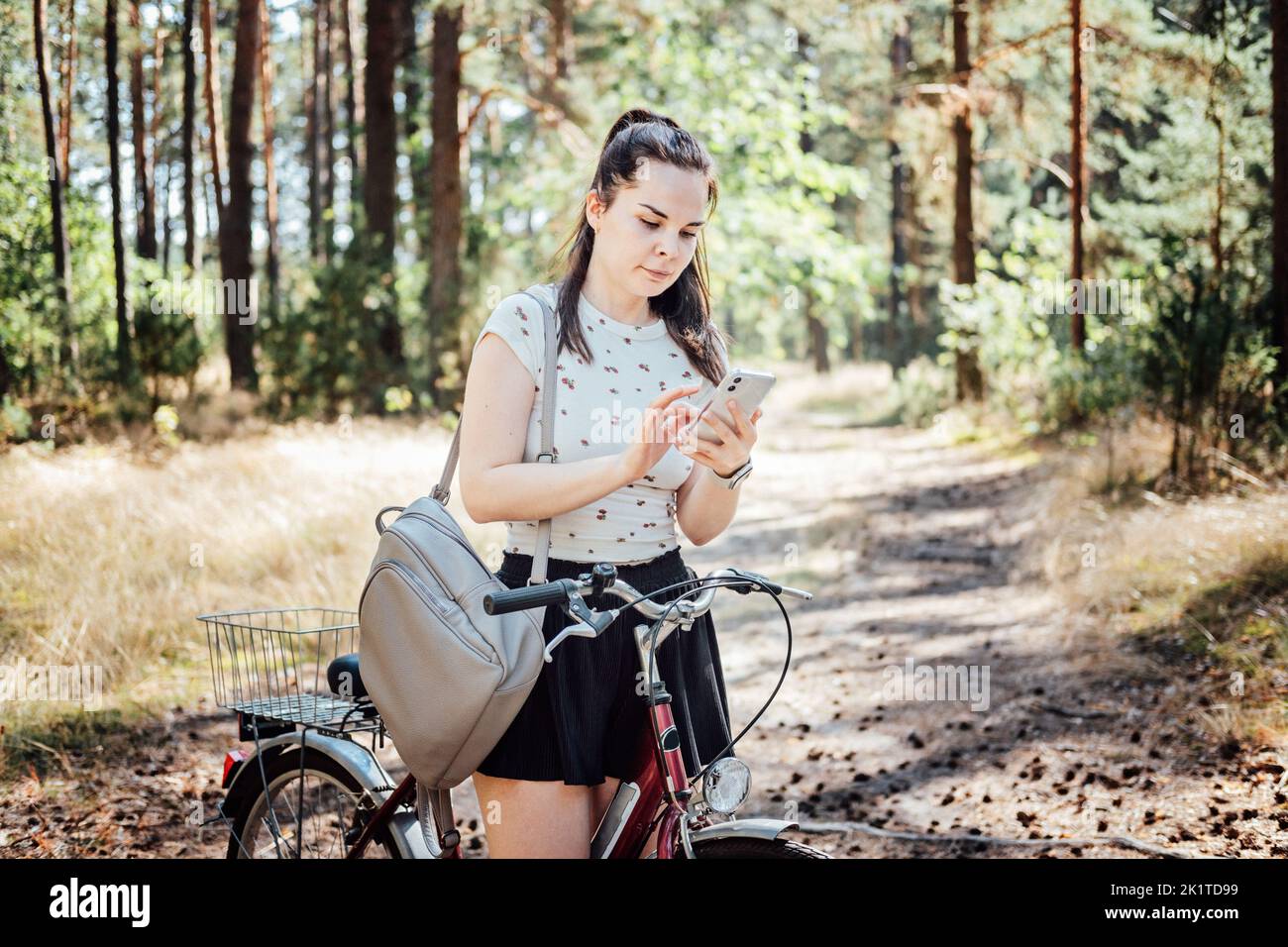 Migliori app per il ciclismo. Bike Tracker. Giovane donna con zaino in bicicletta e guardando in telefono cellulare sullo sfondo della pineta. Ragazza con la bicicletta utilizzando un Foto Stock