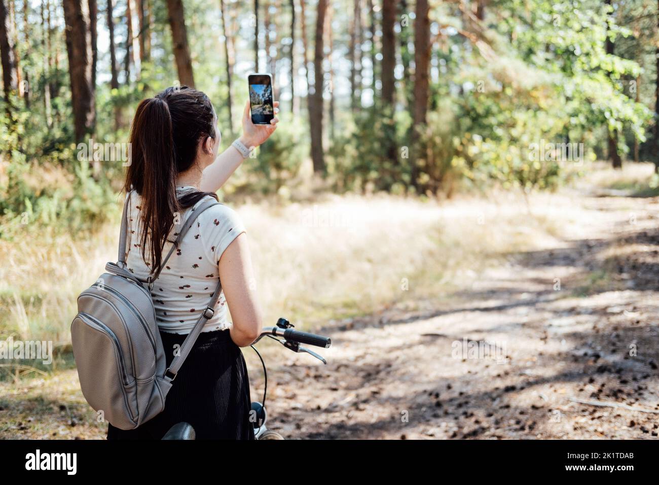 Giovane donna con zaino in bicicletta e scattare foto sul telefono cellulare sullo sfondo della pineta. Ragazza con la bici che scatta foto da telefono fotocamera in Foto Stock