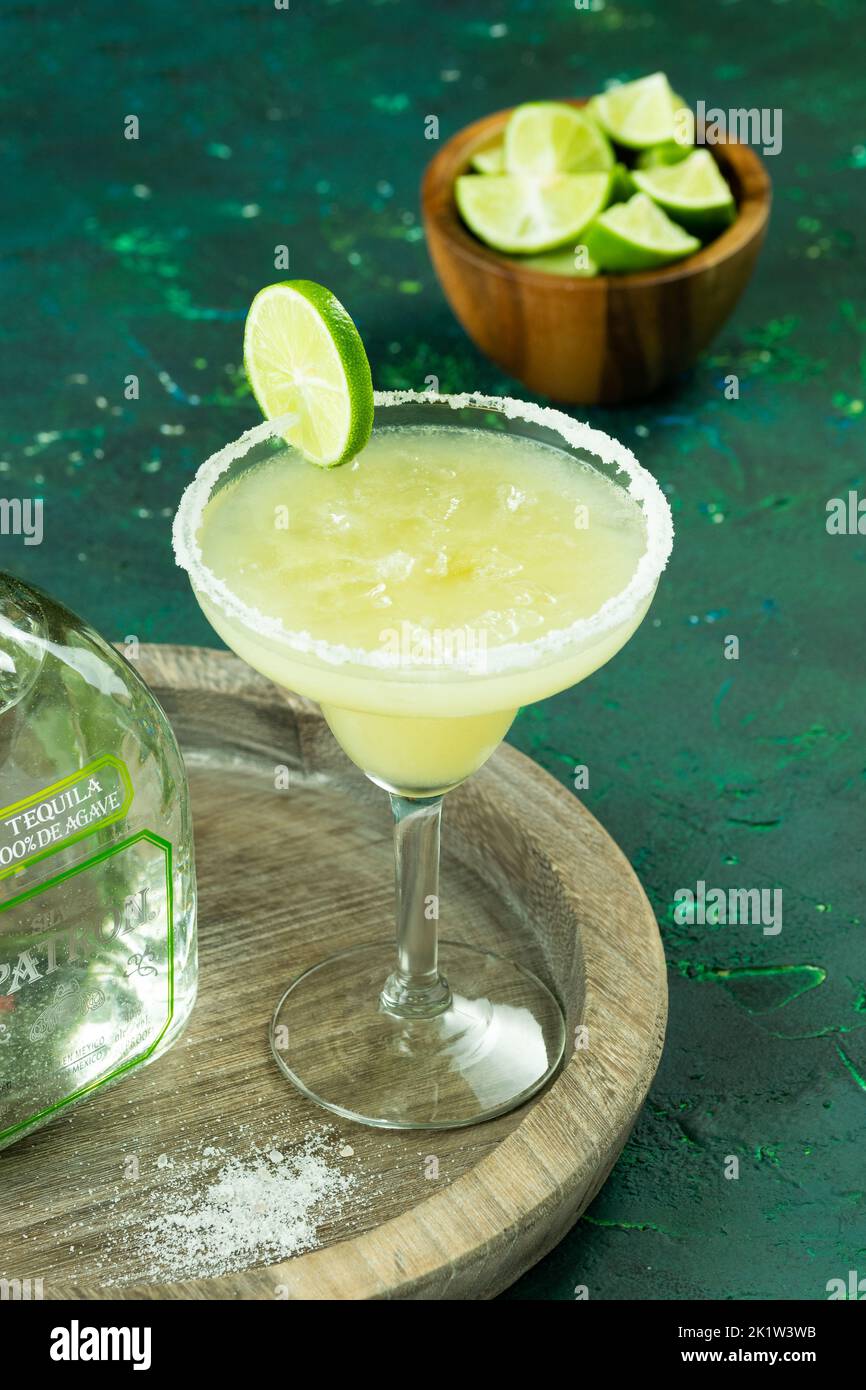 Una verticale di un bicchiere di cocktail Margarita su un piatto di legno accanto ad una bottiglia di tequila e lime a fette Foto Stock