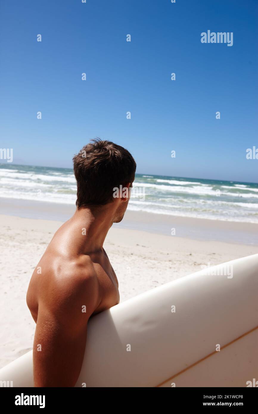 Perfezionare la sua abbronzatura prima di una sessione di surf. La testa e le spalle hanno sparato un giovane in piedi sulla spiaggia con la sua tavola da surf. Foto Stock