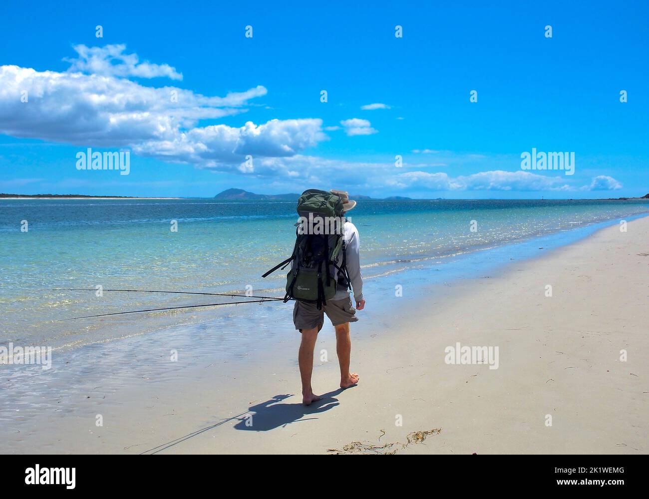 Pescatore solo maschio, vista sul retro, passeggiate lungo la spiaggia di sabbia bianca al bordo dell'acqua. Cielo azzurro estivo e nuvole bianche con colline lontane all'orizzonte. Foto Stock