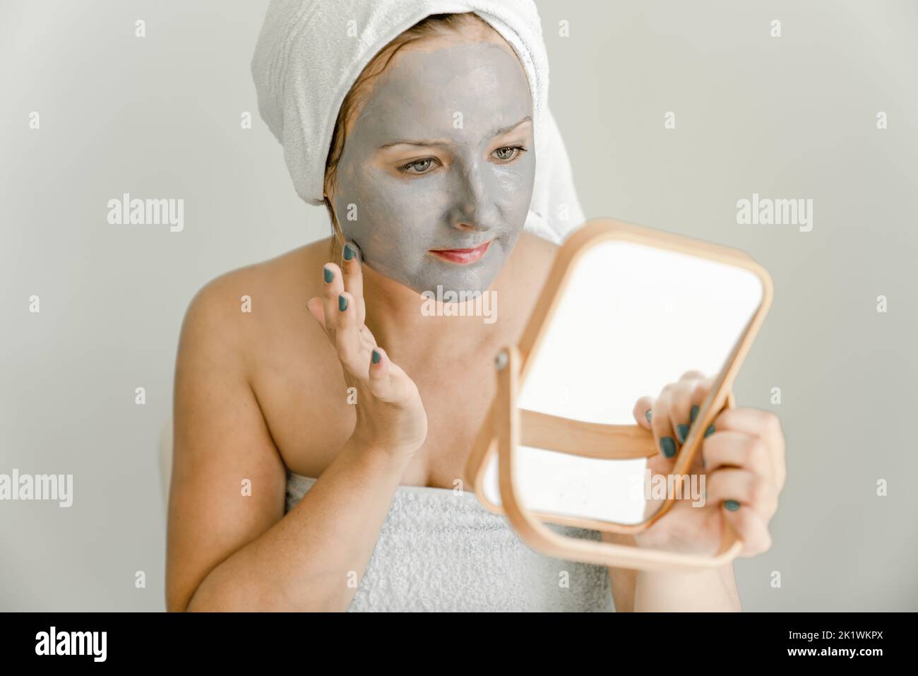 Ragazza avvolta in asciugamano mette maschera di argilla cosmetica grigia sul suo viso mentre guarda in specchio. Primo piano Foto Stock