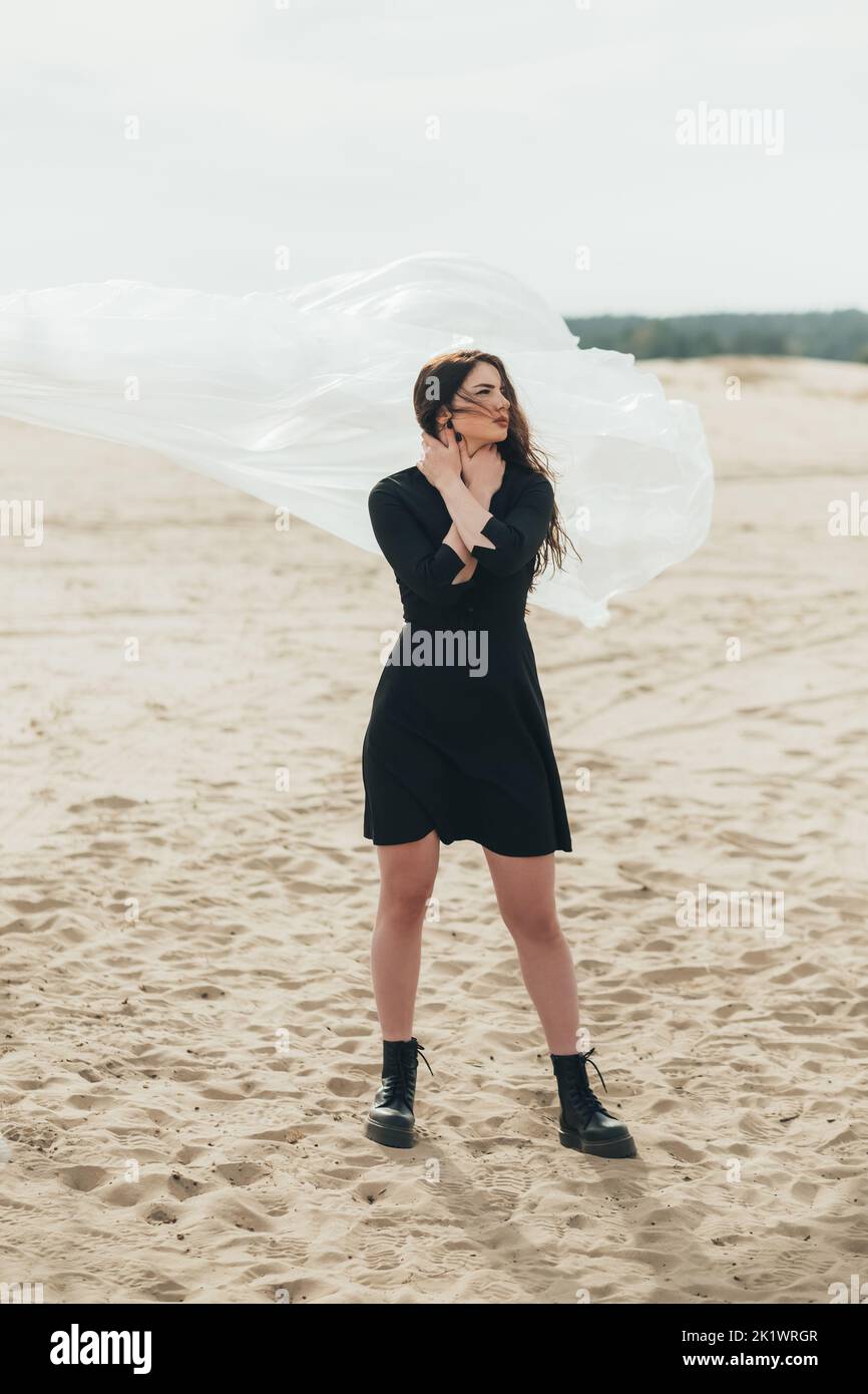 Ritratto di moda. Tenerezza femminile. Autunno malinconia. Donna brunetta pensiva in vestito nero che posa in piedi da solo in vento freddo nel deserto sabbioso con Foto Stock