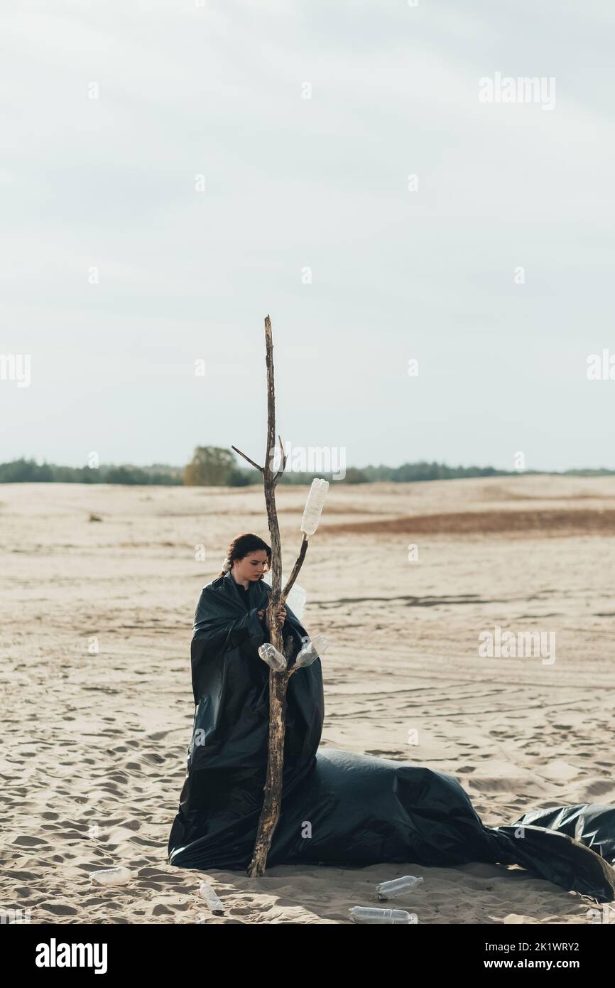 Solitudine femminile. Isolamento sociale. Discriminazione di genere. Donna premurosa depressa che indossa il sacchetto nero della spazzatura in piedi da solo all'albero asciutto in fa fredda Foto Stock