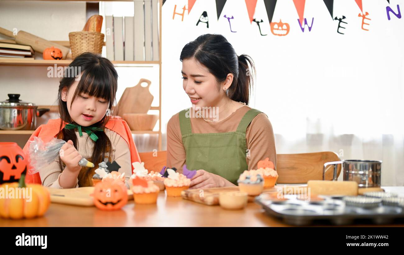 La mamma asiatica gentile e positiva sta insegnando a sua figlia a decorare un cupcake di Halloween. Felice concetto di famiglia Foto Stock