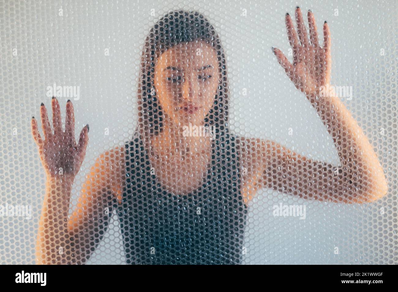 Protezione della donna. Ritratto sfocato. Autoisolamento. Discriminazione femminile. Donna senza speranza nella parte superiore nera intrappolata dietro la texture di pellicola trasparente a bolle Foto Stock