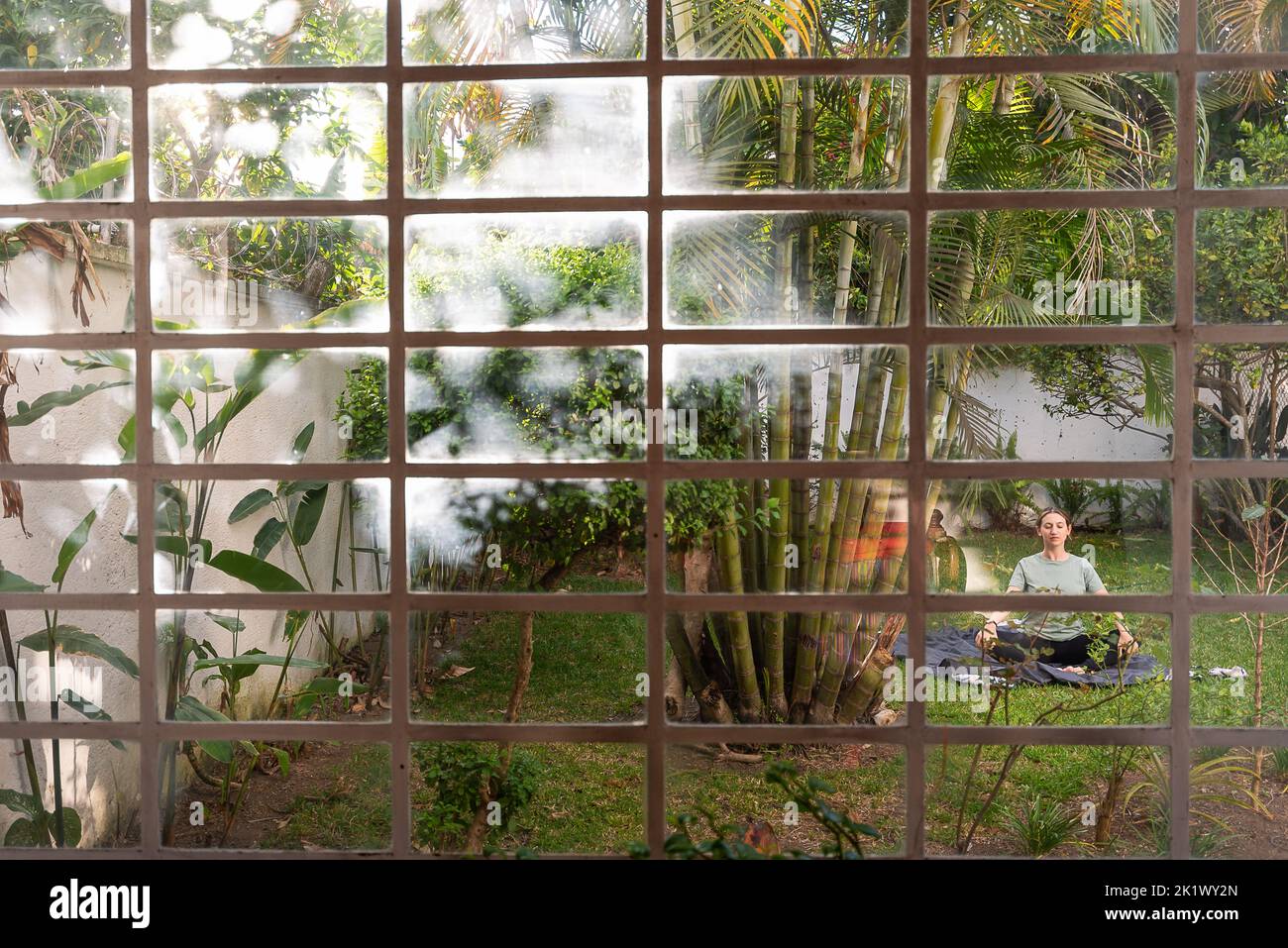 Donna pratica yoga nel cortile circondato da piante e alberi, catturati attraverso la finestra della casa con vibrazioni tropicali e palme Foto Stock