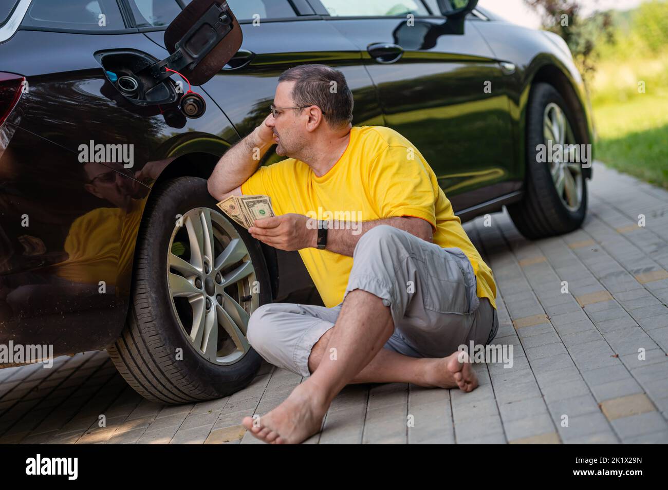 uomo preoccupato con soldi nelle sue mani seduti sul marciapiede accanto ad una macchina vicino ad un serbatoio di gas aperto Foto Stock