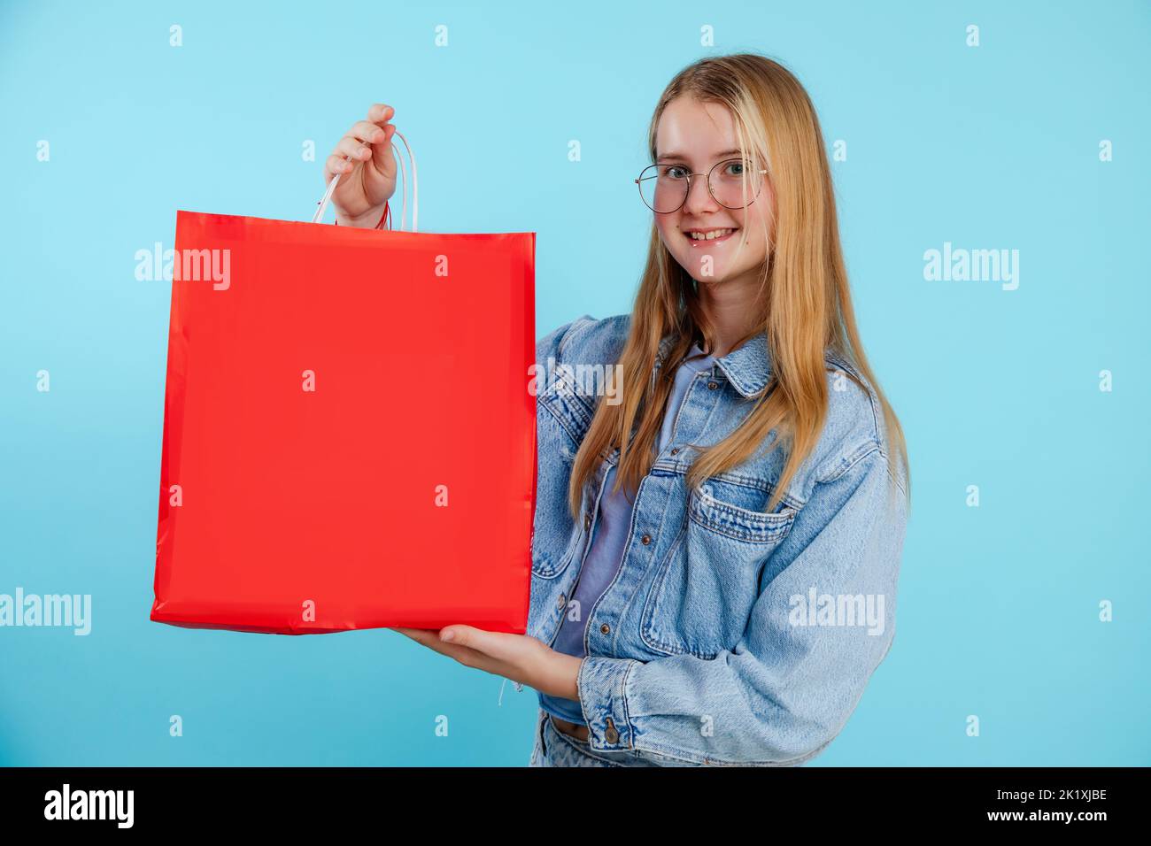 Ragazza giovane bionda sorridente e fiduciosa in occhiali con borsa rossa in mano su sfondo blu. Grandi vendite, shopping Foto Stock