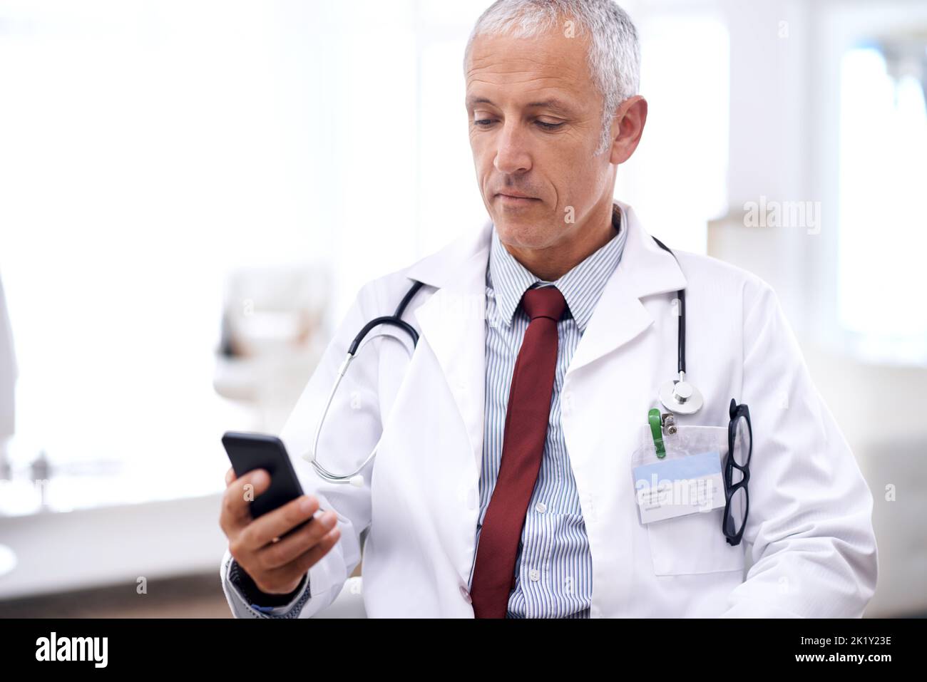 La vostra salute è la mia priorità numero uno: Un medico maturo seduto nel suo ufficio con un cellulare. Foto Stock