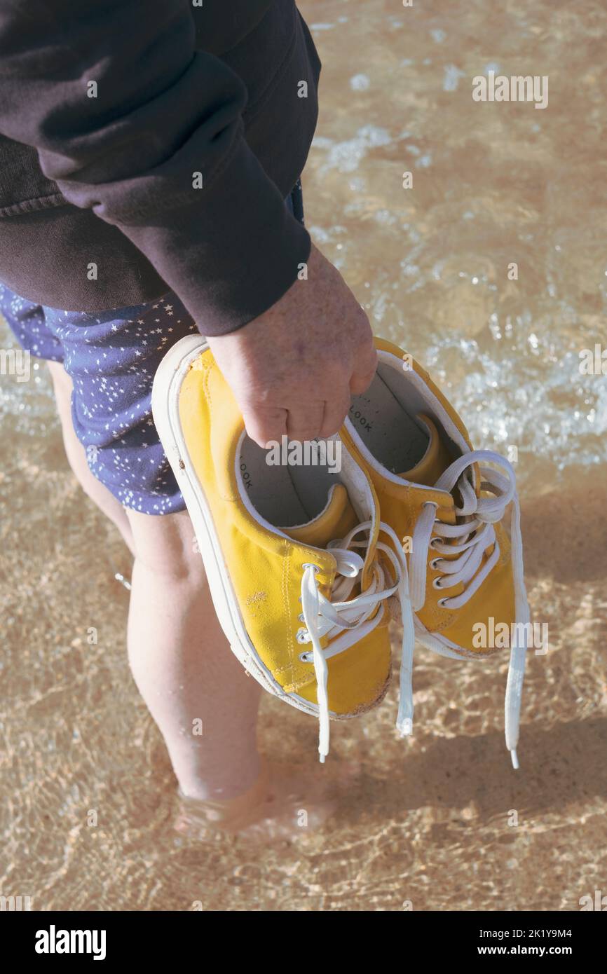 Donna in piedi sul surf sulla spiaggia tenendo scarpe gialle contro lo skyline blu orizzonte Foto Stock