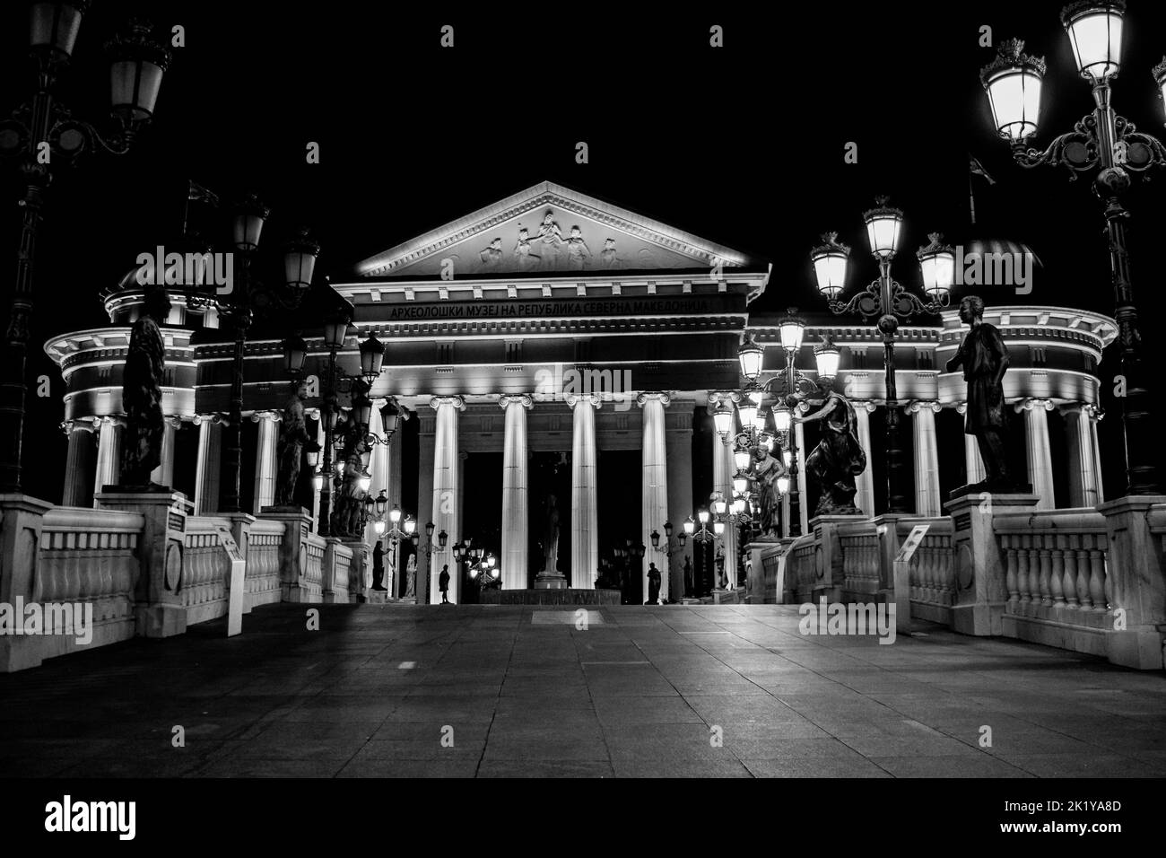 Statue che decorano il Ponte delle civiltà, di fronte al Museo Archeologico di Skopje, Macedonia settentrionale. Di notte con lampioni che illuminano il ponte. Foto Stock