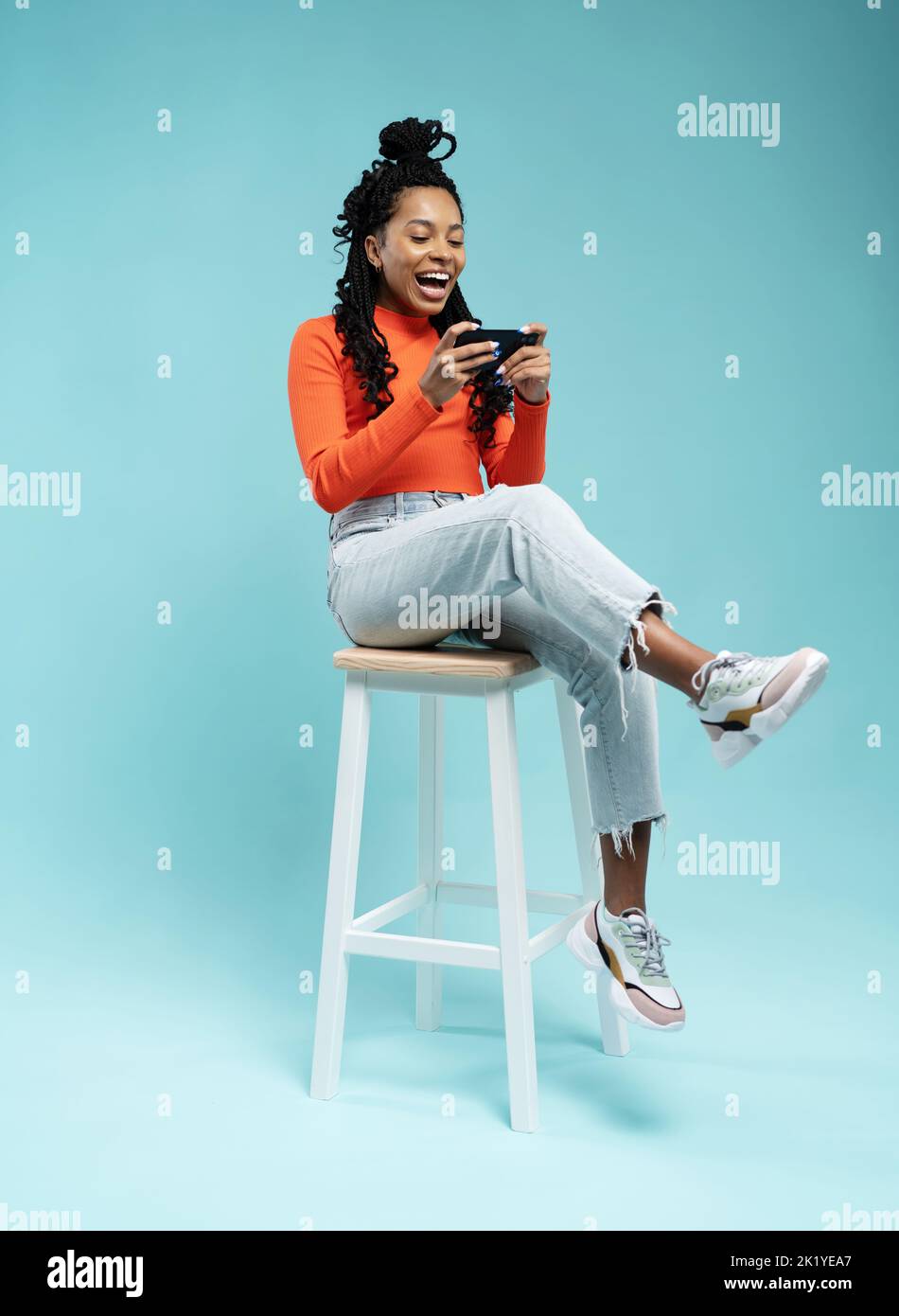 Ritratto di una giovane donna eccitata seduta su uno sgabello e giocare a giochi su un telefono cellulare isolato su sfondo blu Foto Stock