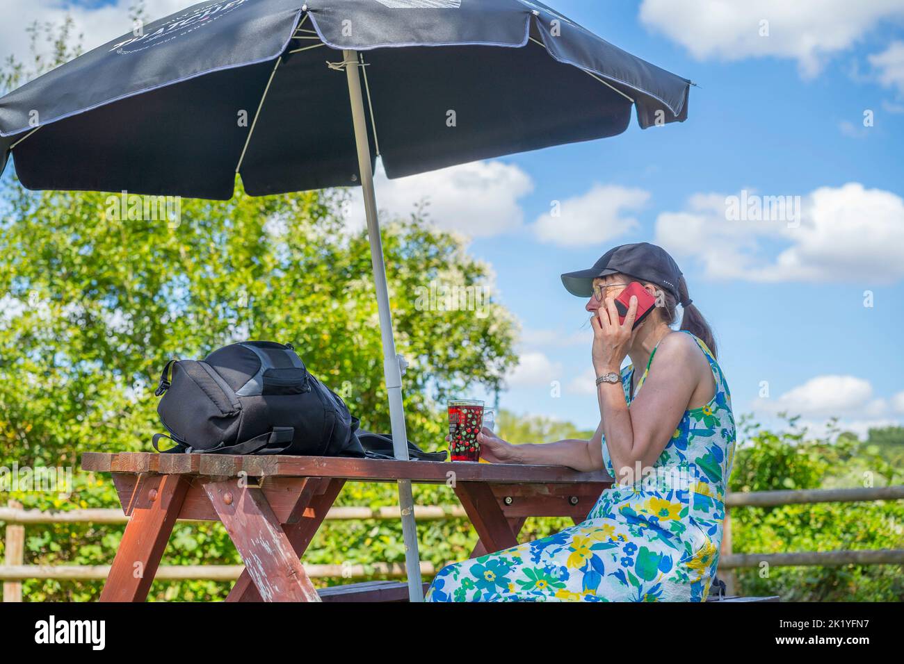 Vista laterale di una donna seduta a bere in un pub giardino in una giornata di sole mentre parla sul suo telefono cellulare. La donna si rifugia sotto un giardino di sole brolly a pr Foto Stock