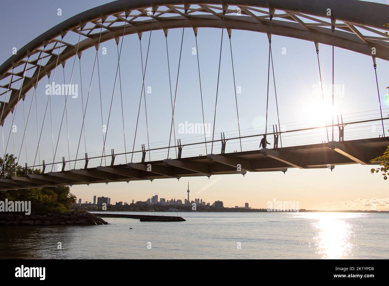 Il sole della mattina presto splende attraverso il Ponte dell'Arco di Humber Bay mentre una persona cammina. La CN Tower e lo skyline di Toronto sono visibili sullo sfondo. Foto Stock