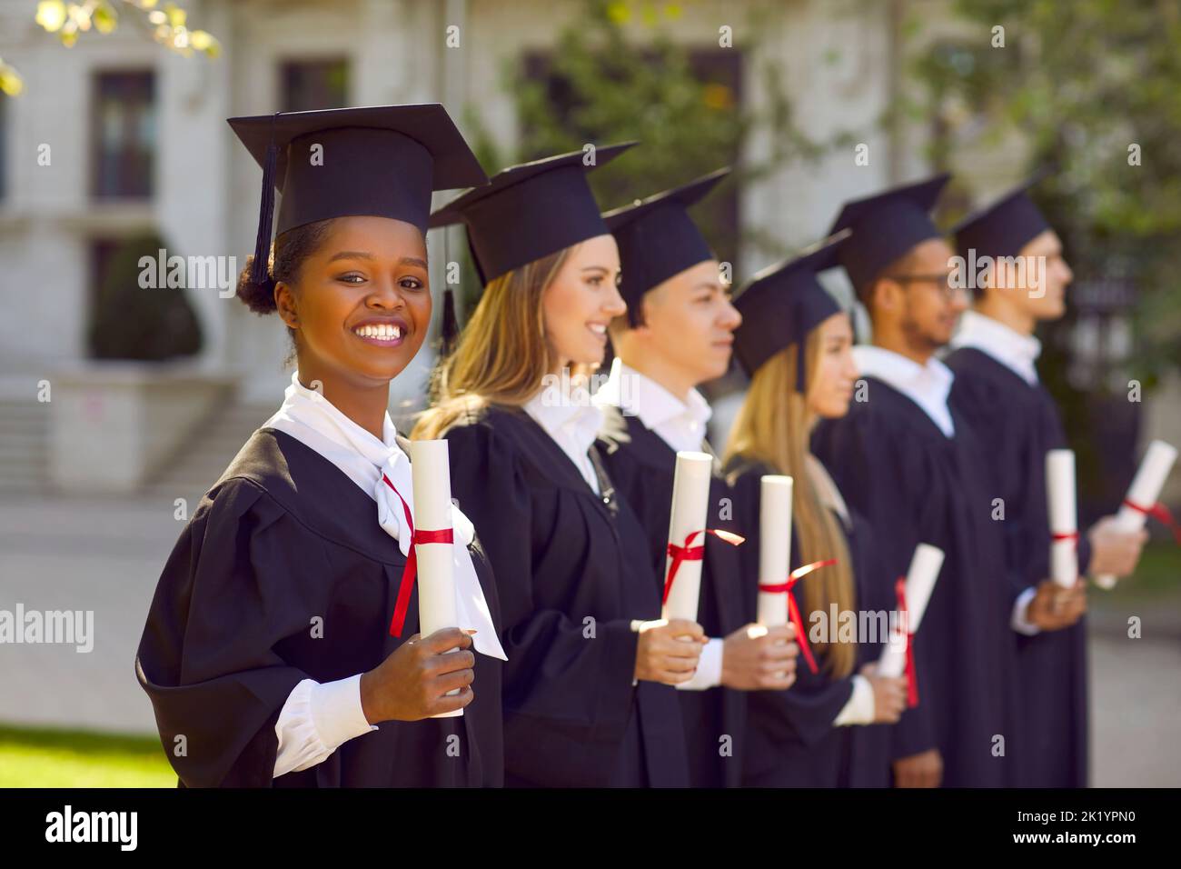 Giovane studentessa afroamericana in abito di laurea e cappello sorride si alza con i laureati Foto Stock