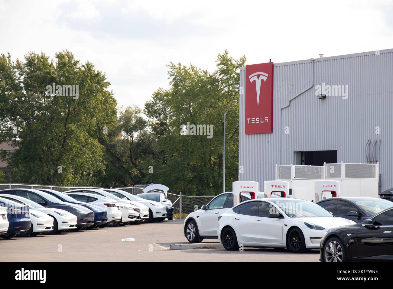 Il logo Tesla è visibile sul lato di una concessionaria Tesla e di una stazione di ricarica Supercharger, piena di auto in carica e di veicoli nuovi nel parcheggio. Foto Stock