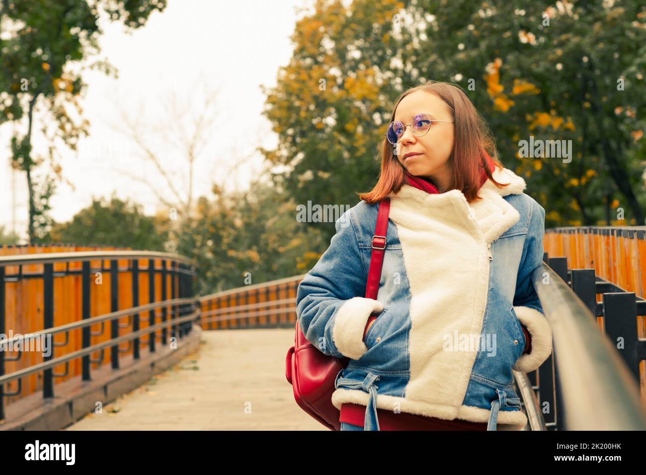 Giovane bella donna in occhiali con capelli corti in giacca calda denim guardando lontano, in piedi sul ponte pedonale in tempo fresco Foto Stock